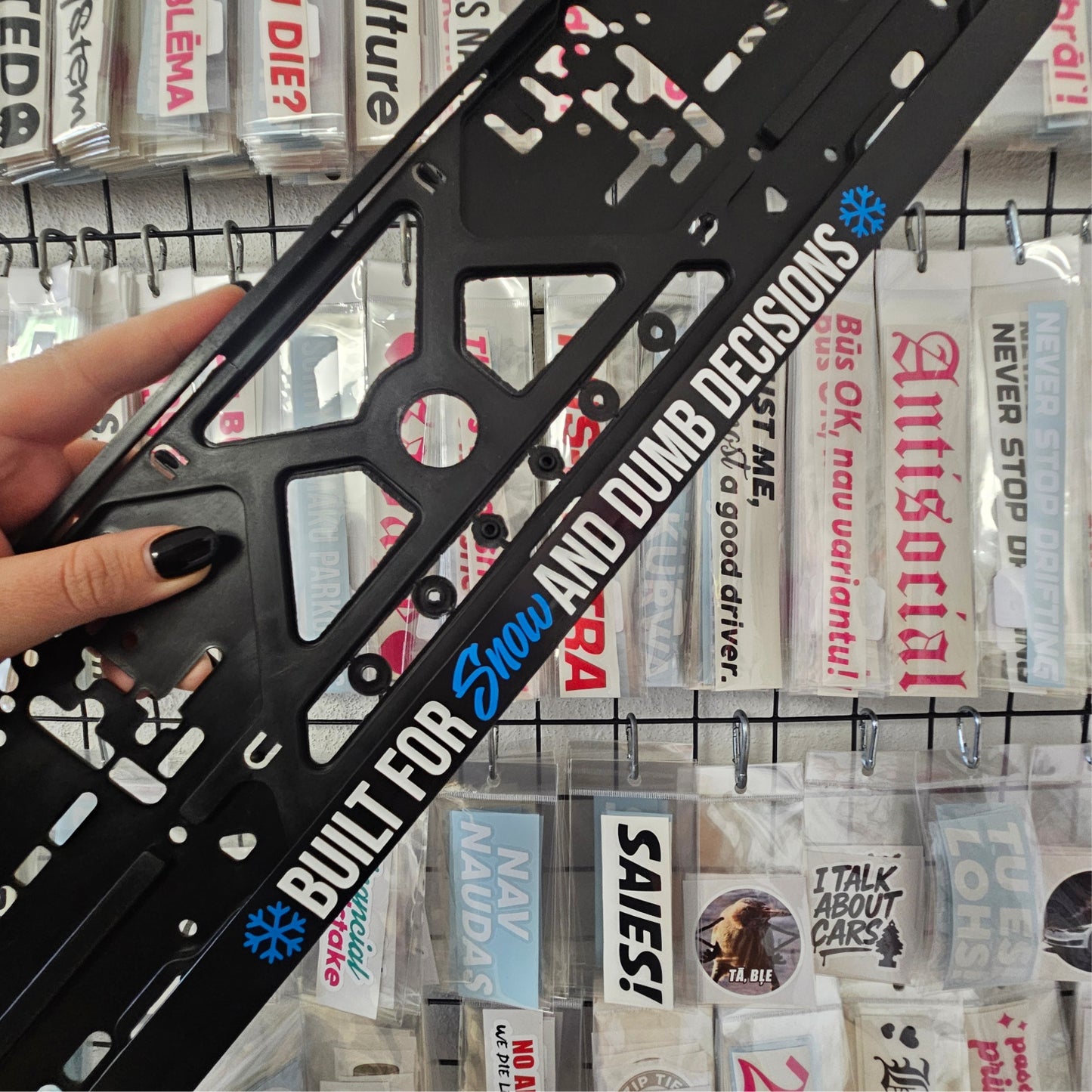 Black license plate frame with text and graphics held by a hand, with other license plate frames in the background.