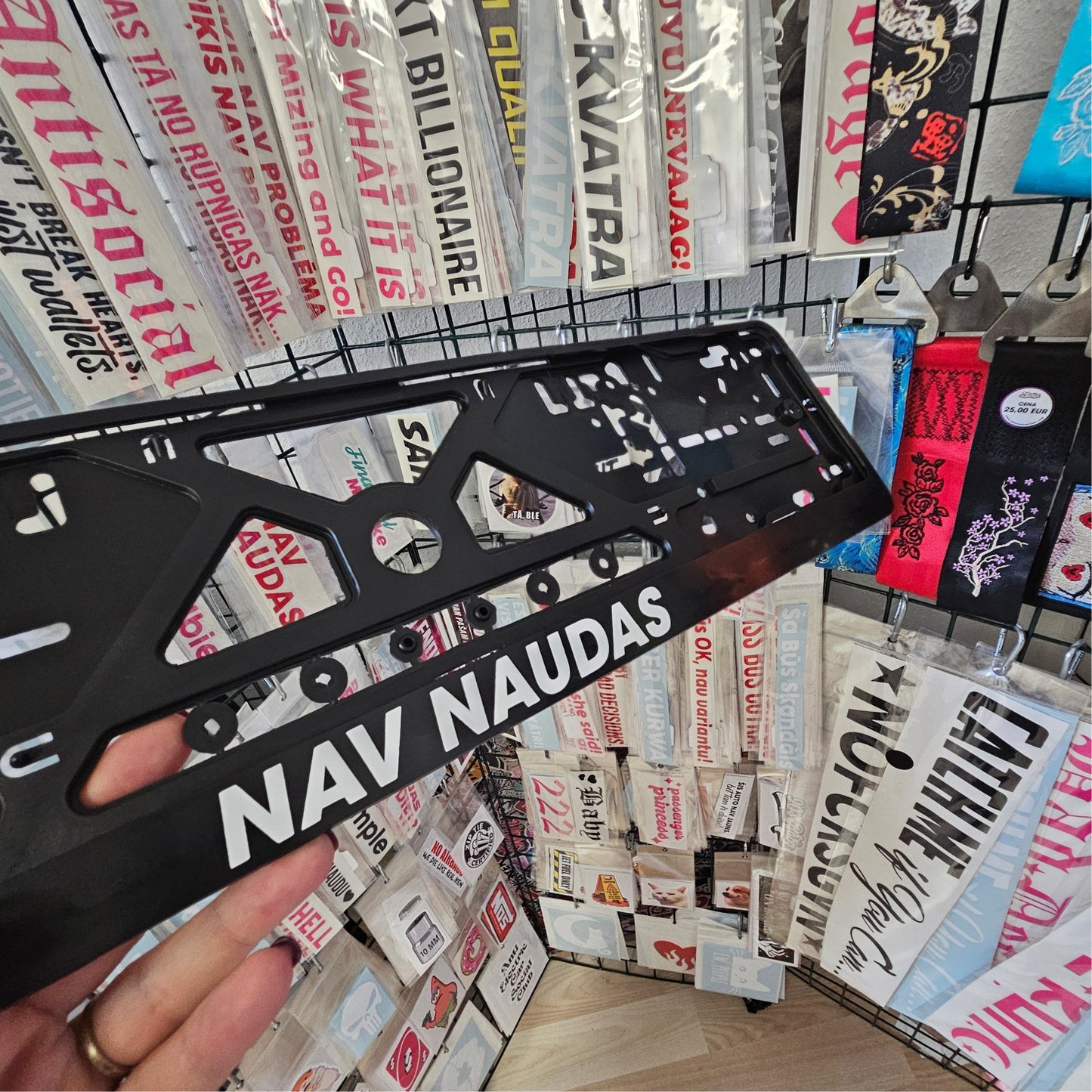 Black license plate frame with 'NAV NAUDAS' text held in front of various printed products.