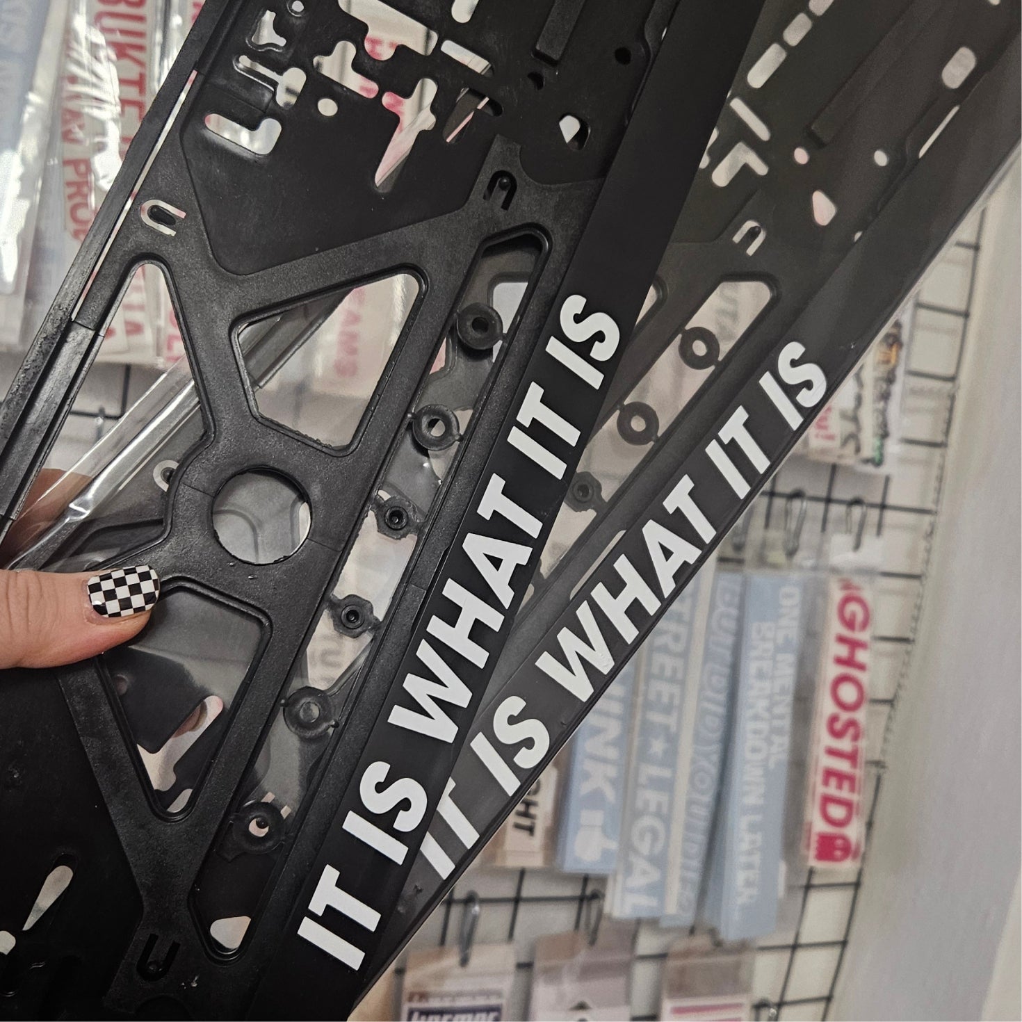 Black metal bracket with 'IT IS WHAT IT IS' text held by a hand with checkered nail polish.