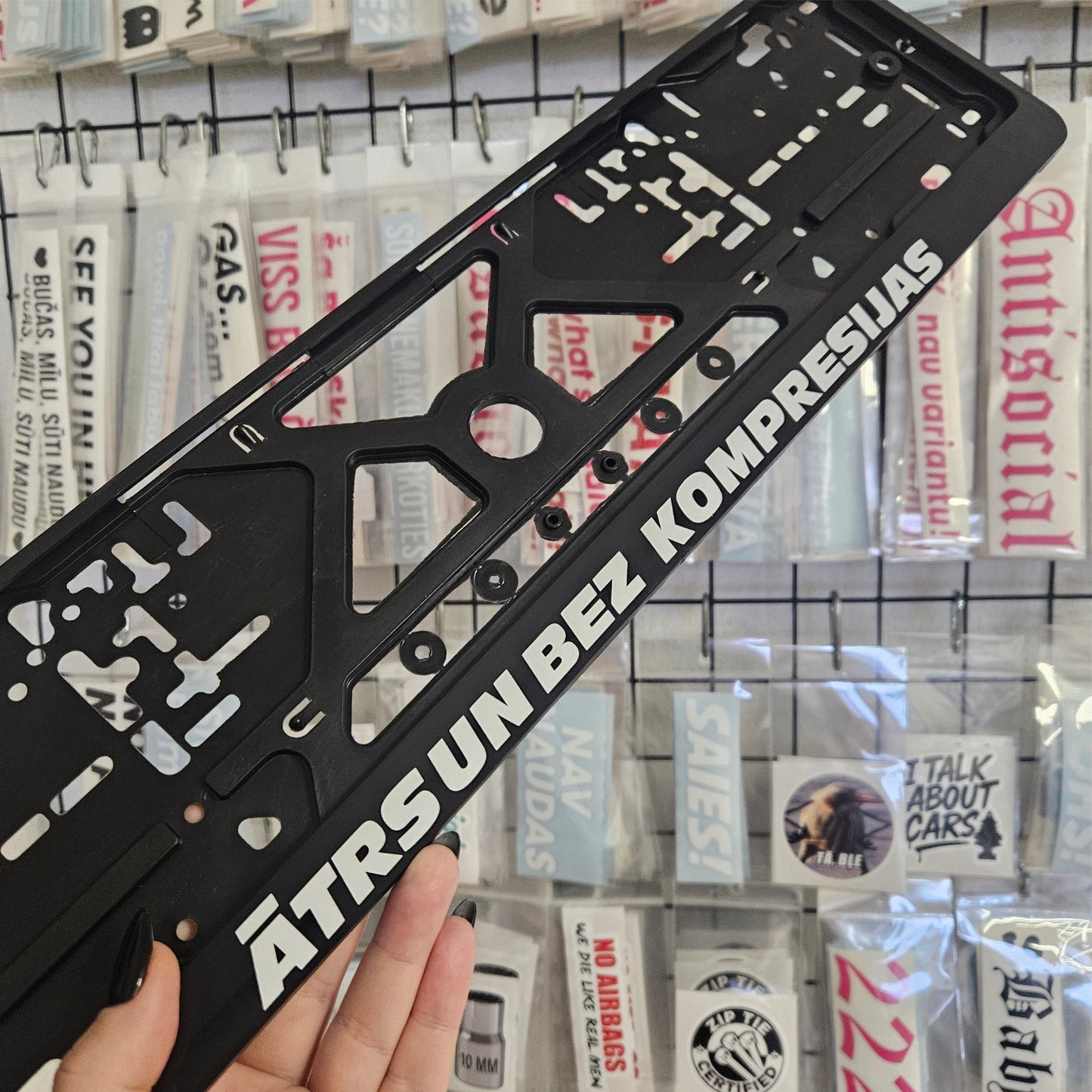 Black license plate frame with white text held in front of a display of stickers.