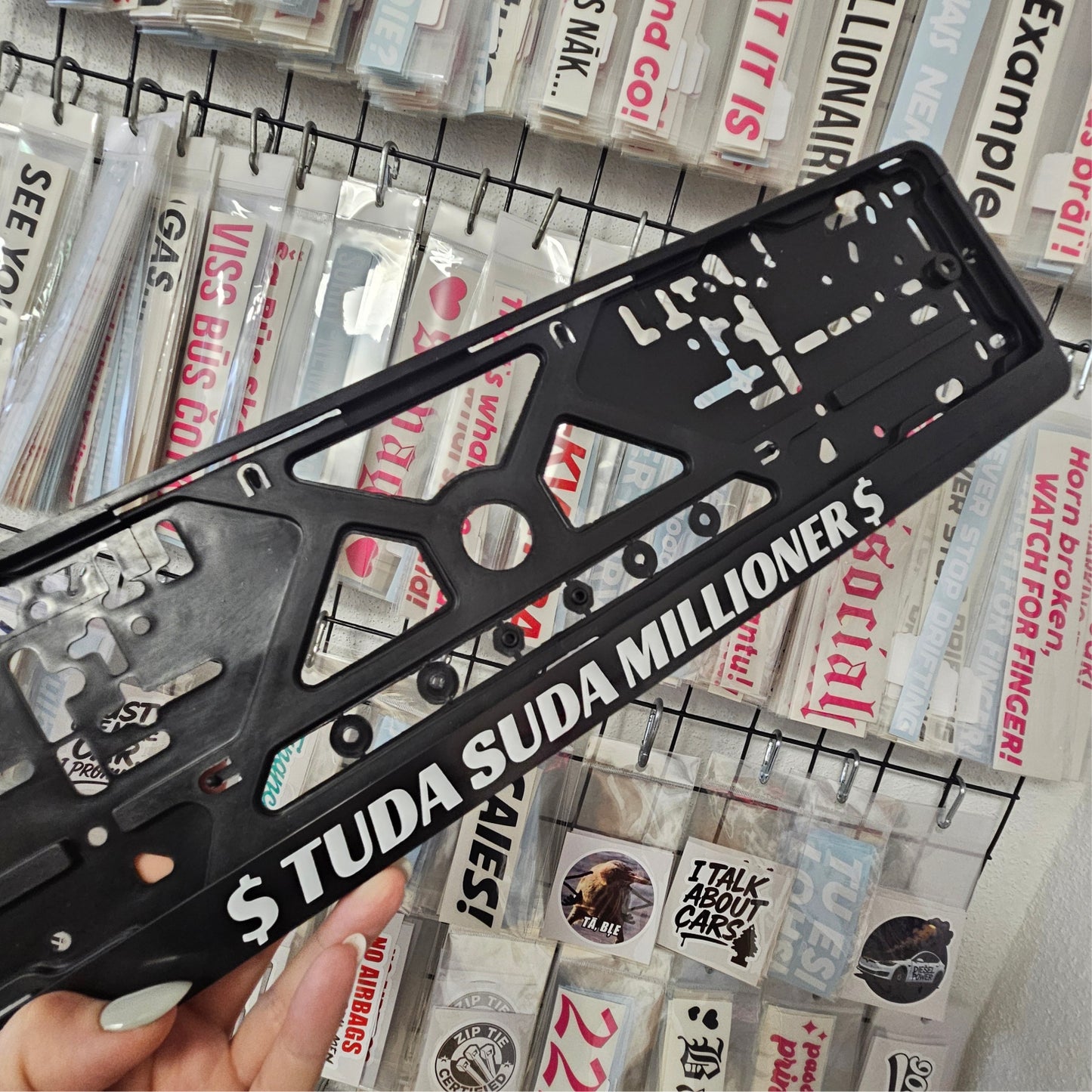 Black license plate frame with 'S TUDA SUDA MILLIONERS' text held over a wall of various license plates.