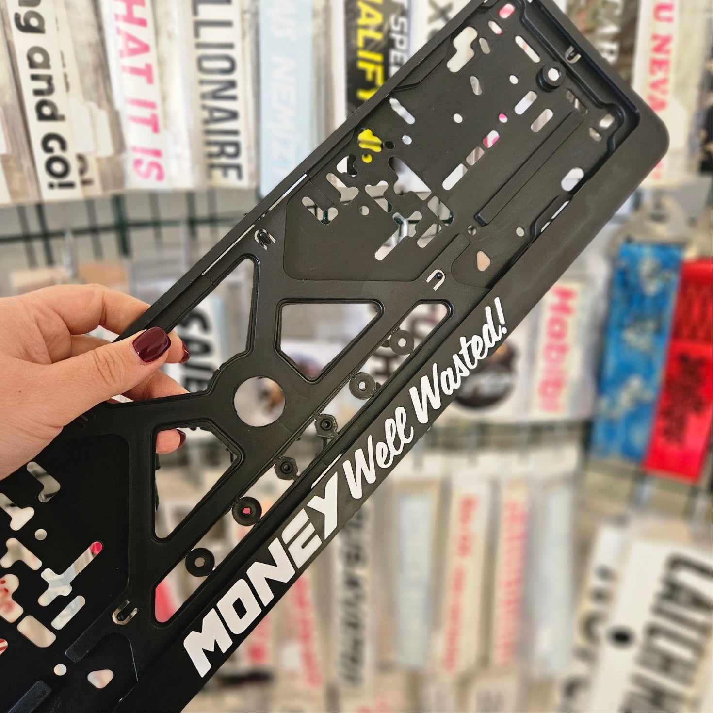 Black license plate frame with 'Money Well Wasted!' text held in front of a bookshelf.