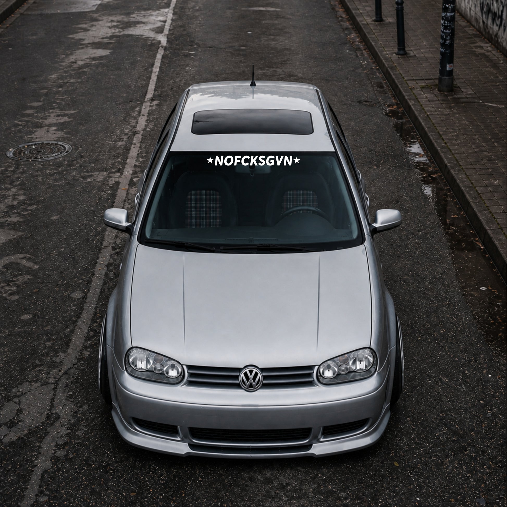 Silver Volkswagen car with a distinctive sticker on the windshield, parked on an urban street.