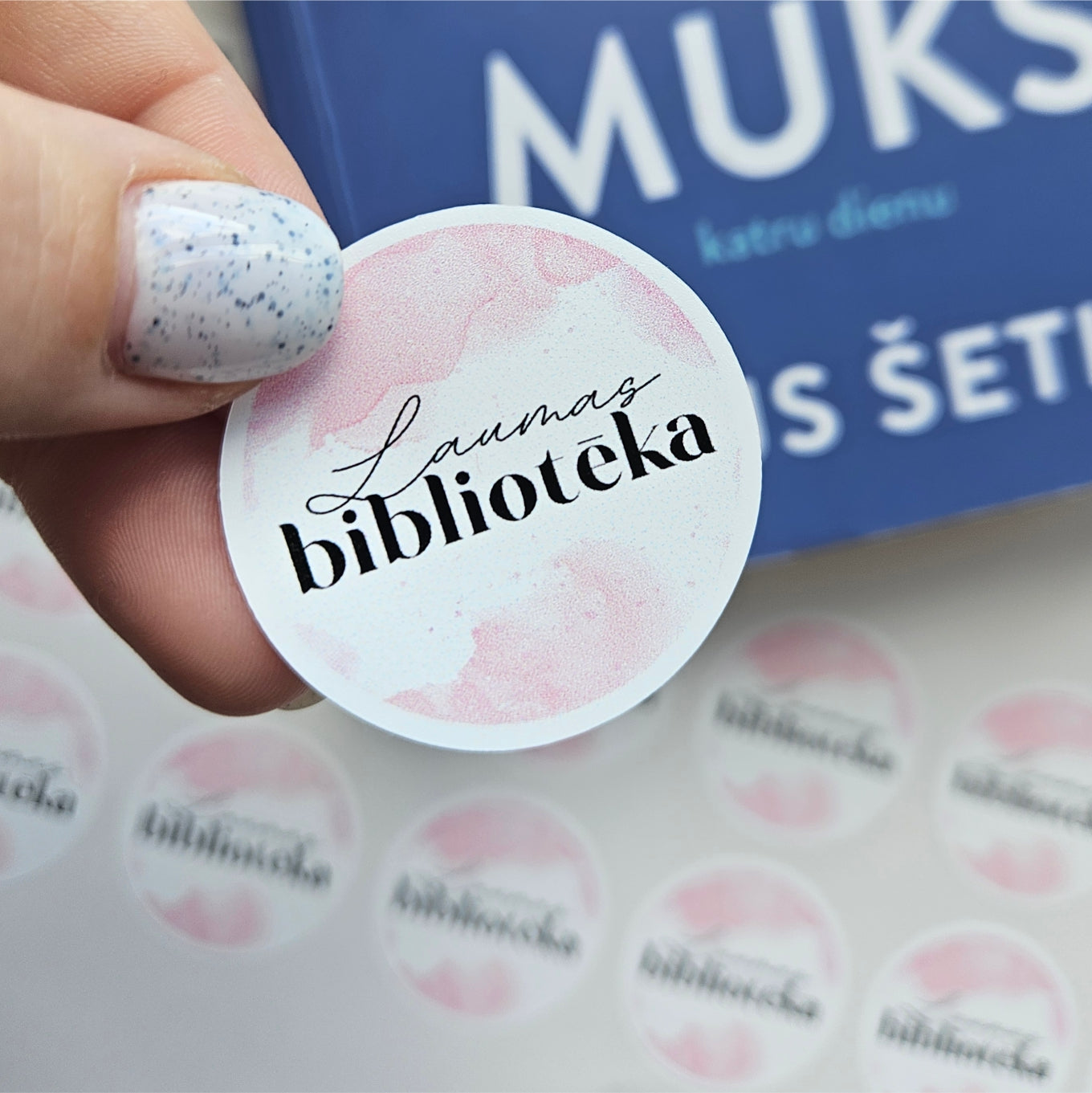 Round sticker with 'Laumas biblioteka' text held by a hand, with a blue book in the background.