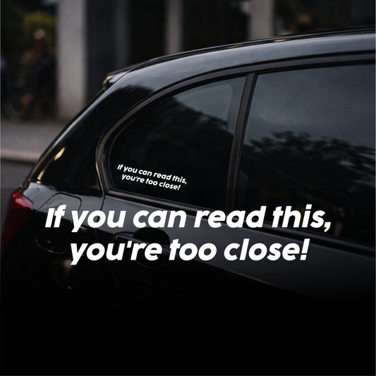 Car window with a humorous sticker reading 'If you can read this, you're too close!' on a dark background.