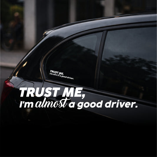Car window with a humorous decal reading 'Trust me, I'm almost a good driver.'