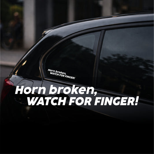 Car window with a humorous sticker warning 'Horn broken, watch for finger!' on a dark background.
