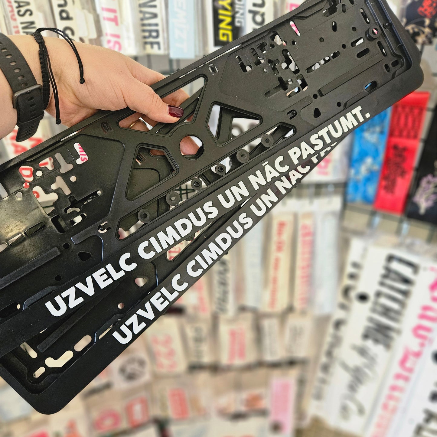 Black license plate frame with text held by a hand against a blurred store background