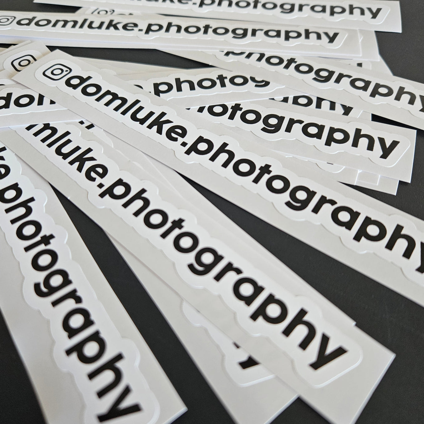 Multiple 'domluke.photography' stickers on a dark surface