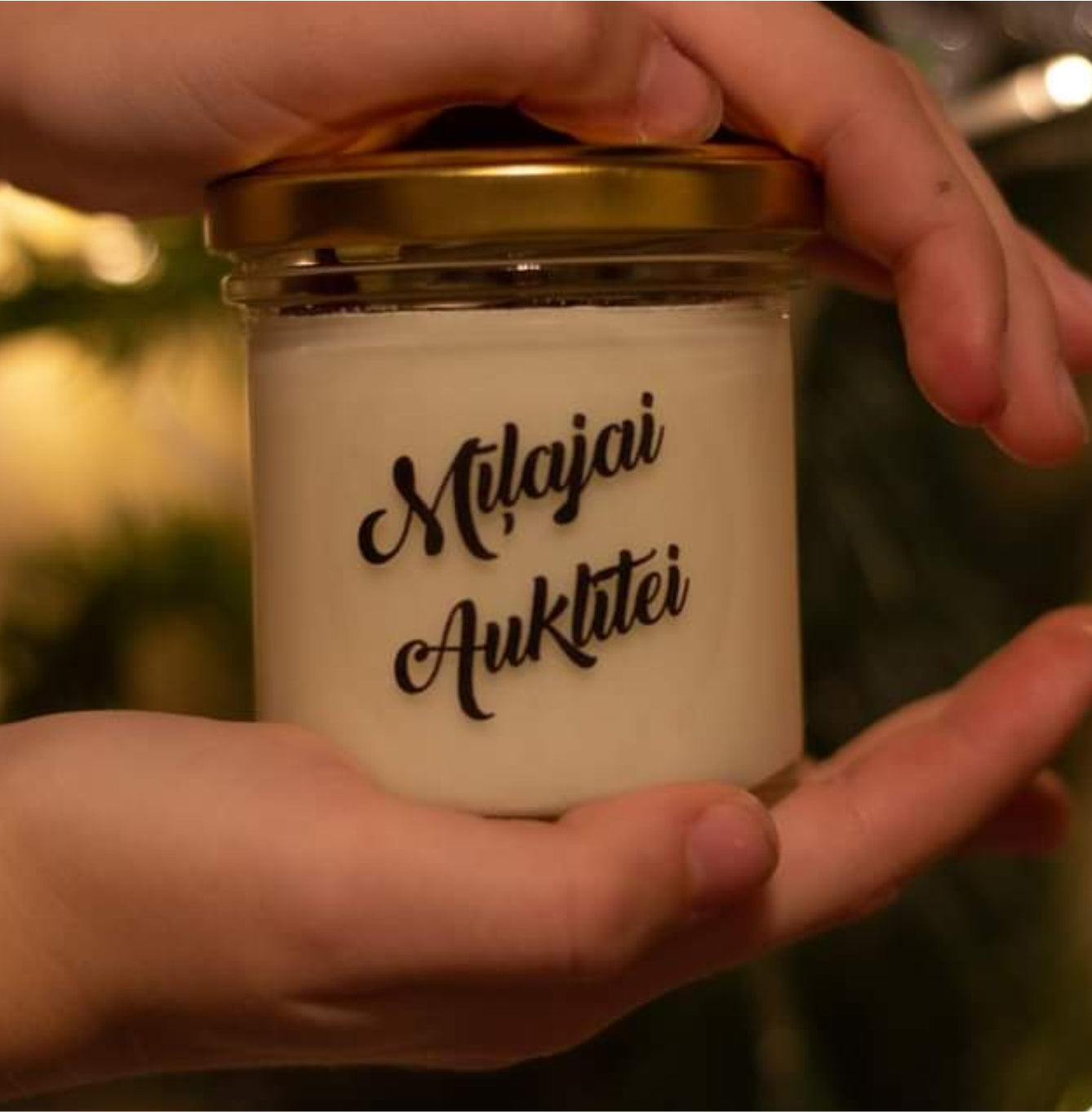 Candle jar with 'Milájai Auklátié' text held by a hand against a blurred background